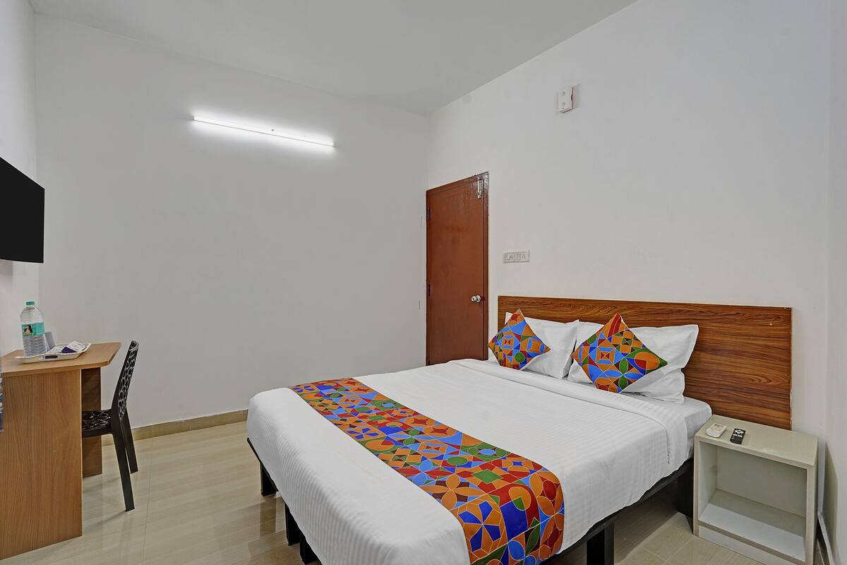 A hotel room with a bed, a wooden nightstand, a desk with a chair, and a television mounted on the wall.