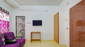 A room with a purple sofa, a small wooden table, a TV mounted on the wall, and a wooden door. - Fabexpress Ss Residency (Chennai)