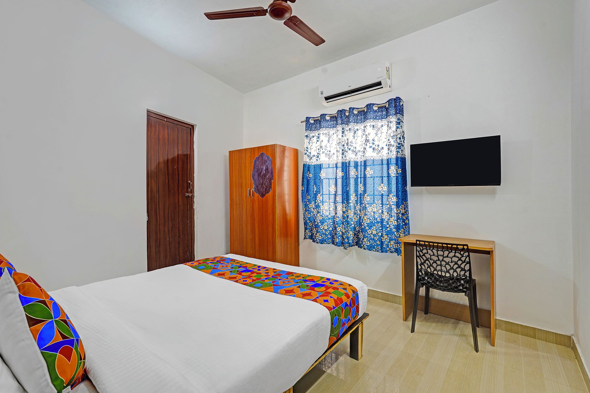A bedroom with a bed, a wooden wardrobe, a desk with a chair, a ceiling fan, an air conditioning unit, and a TV mounted on the wall.