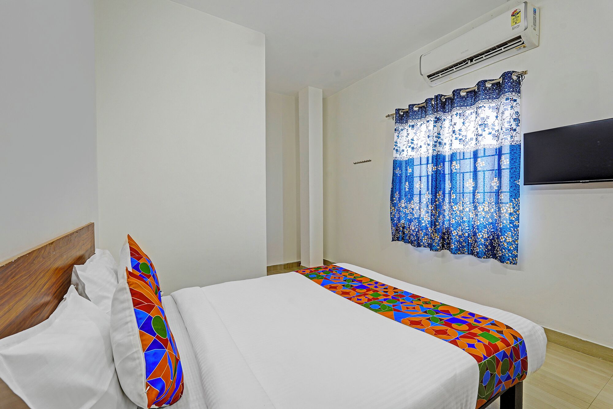 A small, clean room with a bed, a window with a blue floral curtain, and an air conditioning unit.