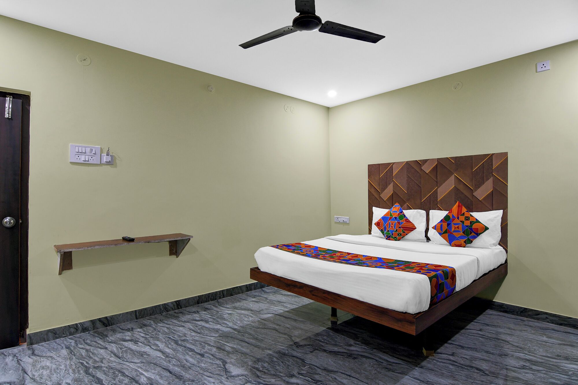 A hotel room with a bed, a ceiling fan, a bedside table, and a wall-mounted shelf.