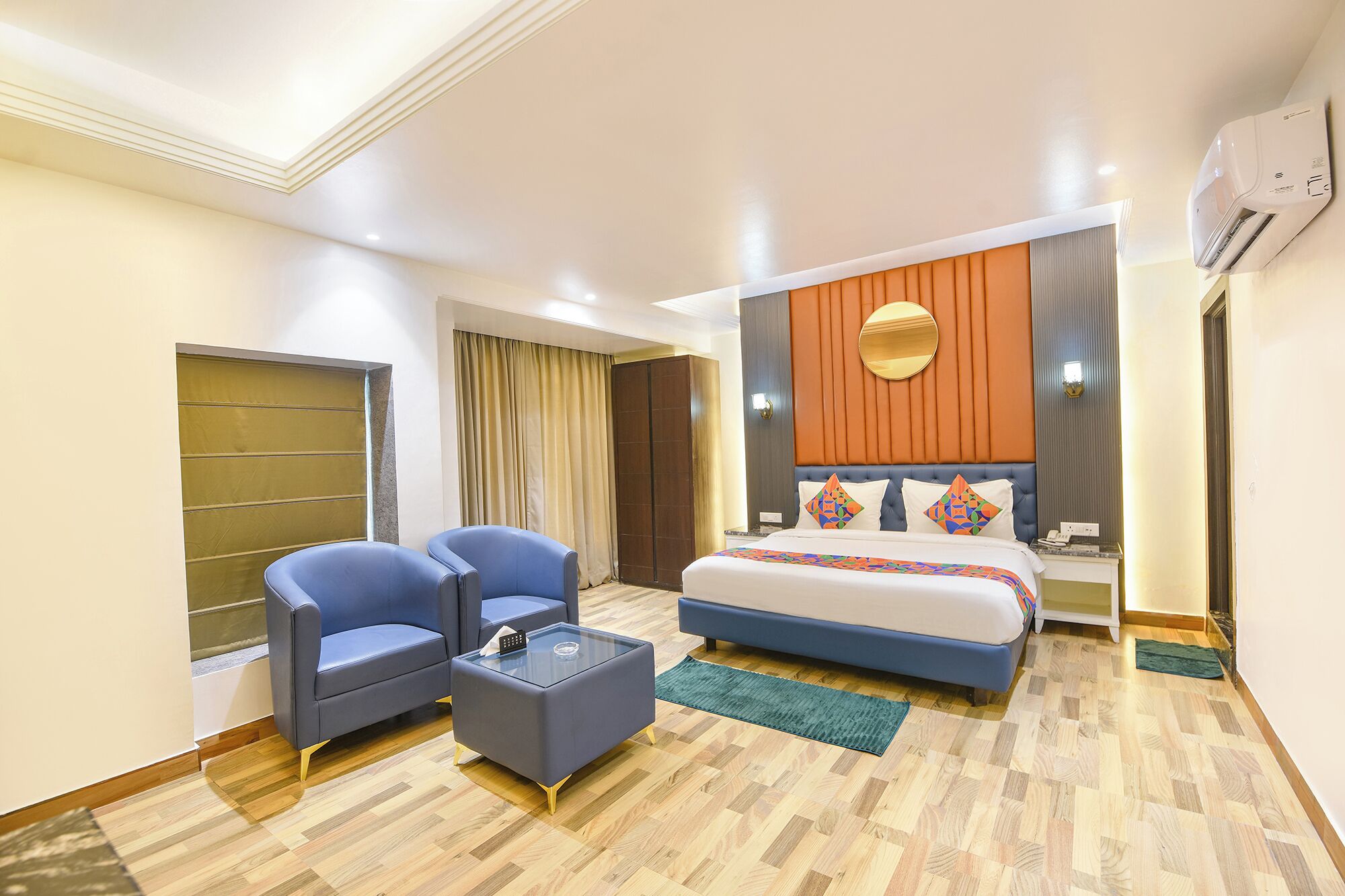 A hotel room with a bed, two armchairs, a small table, and a wall-mounted air conditioner.
