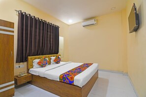 A hotel room with a wooden bed, white bedding, a colorful geometric throw, a brown curtain, a wooden wardrobe, a bedside table with a tray, and an air conditioning unit on the wall. - Via Philips (Kolkata)