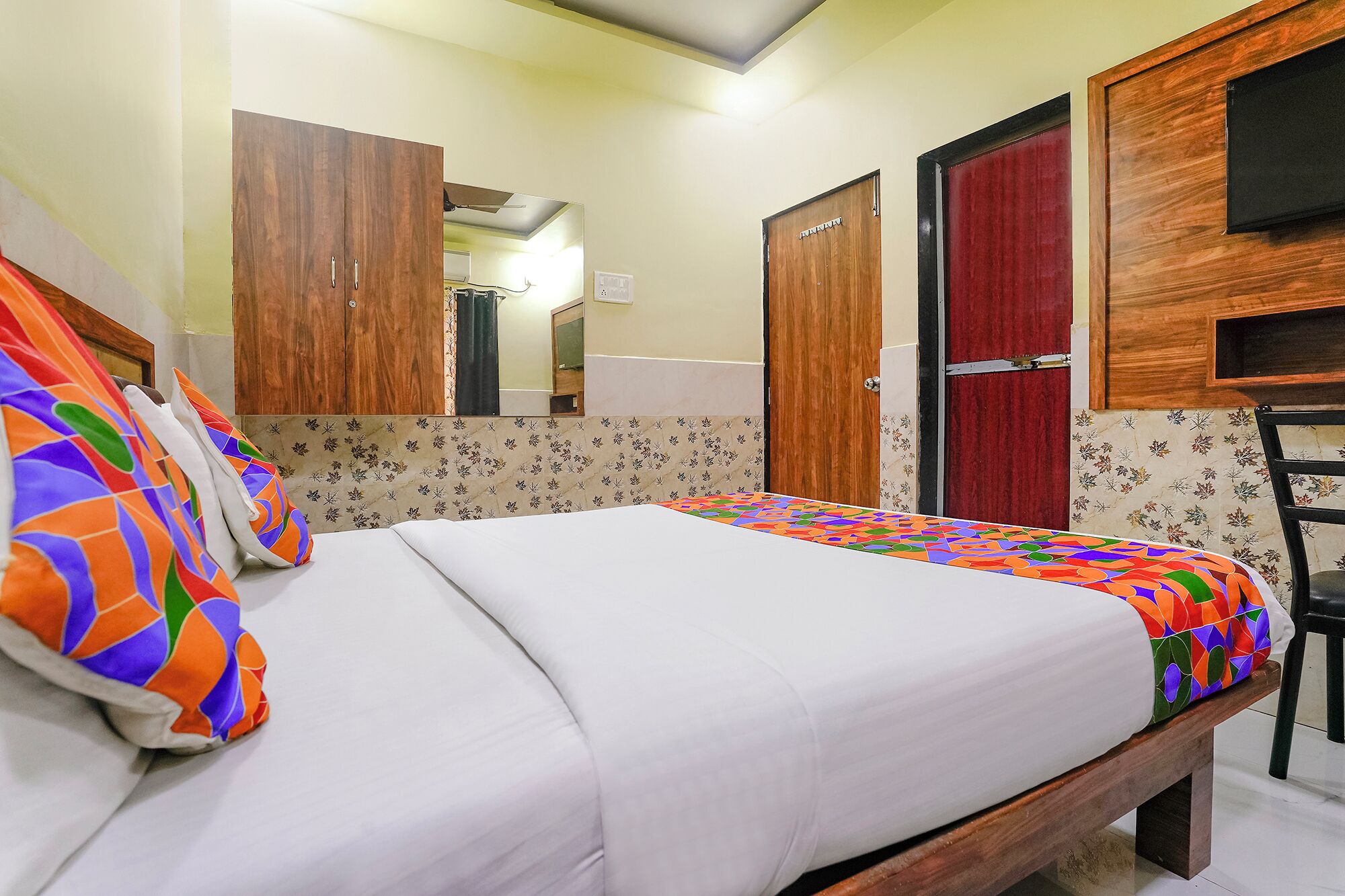 Una cama bien hecha con almohadas coloridas, un armario de madera, una puerta roja y un espejo.