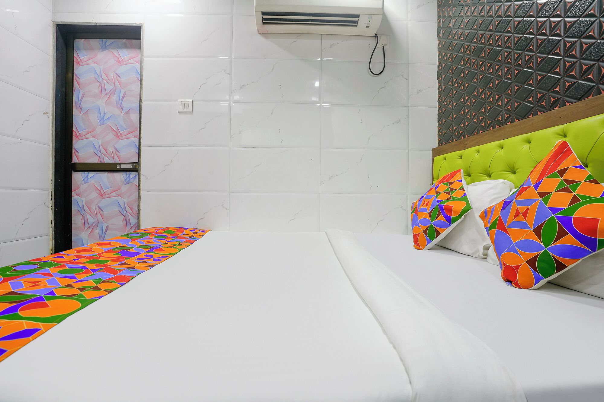 A hotel room with a bed, colorful pillows, a white headboard, a wall air conditioner, and a geometric-patterned door.