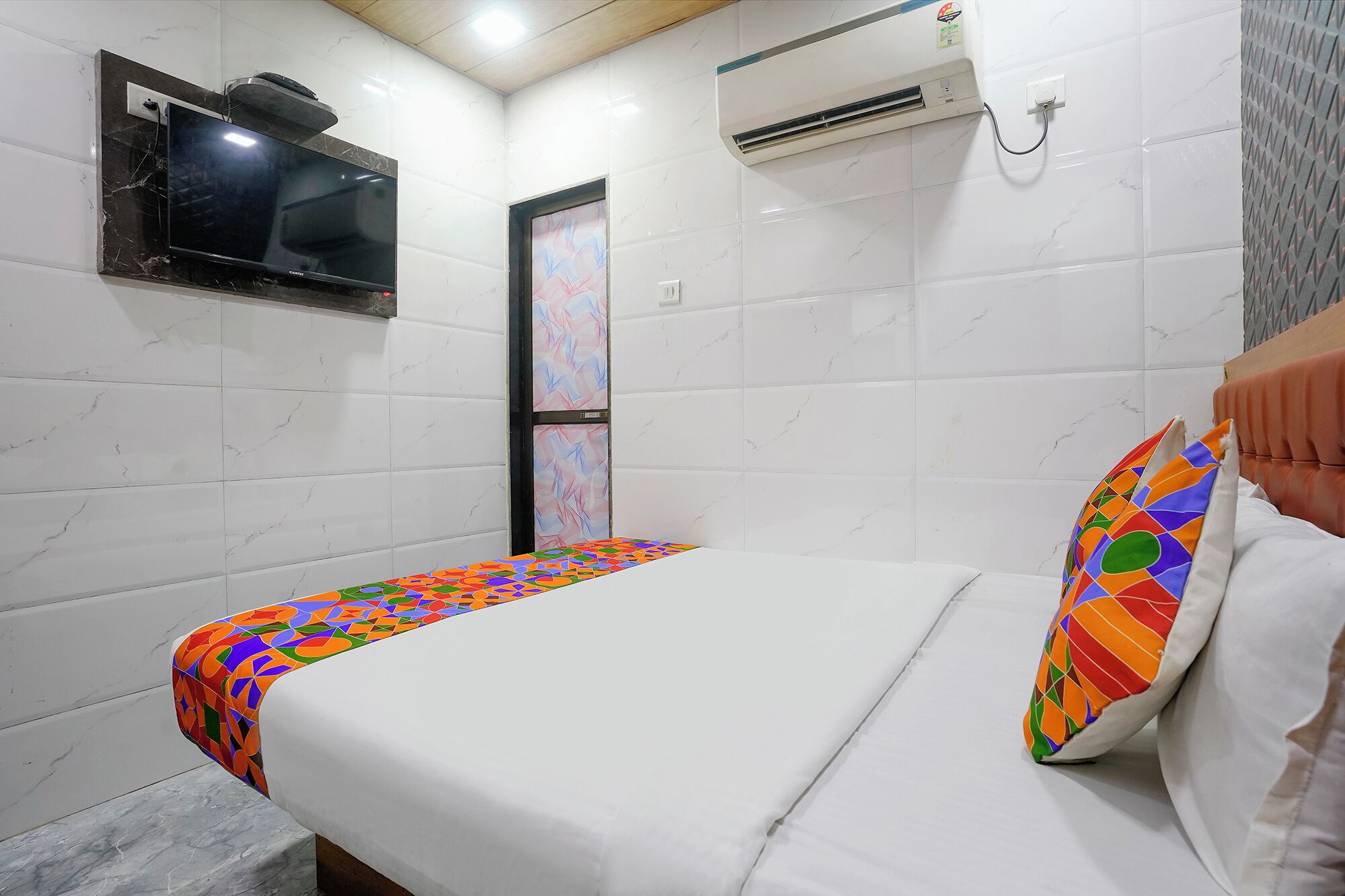A hotel room with a bed, a TV mounted on the wall, and an air conditioning unit.