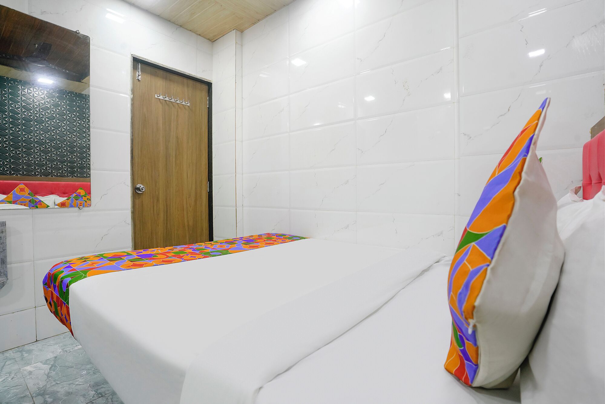 Ein kleines Zimmer mit einem Einzelbett, einer Holztür, weißen Fliesenwänden und einer farbenfrohen Bettdecke.