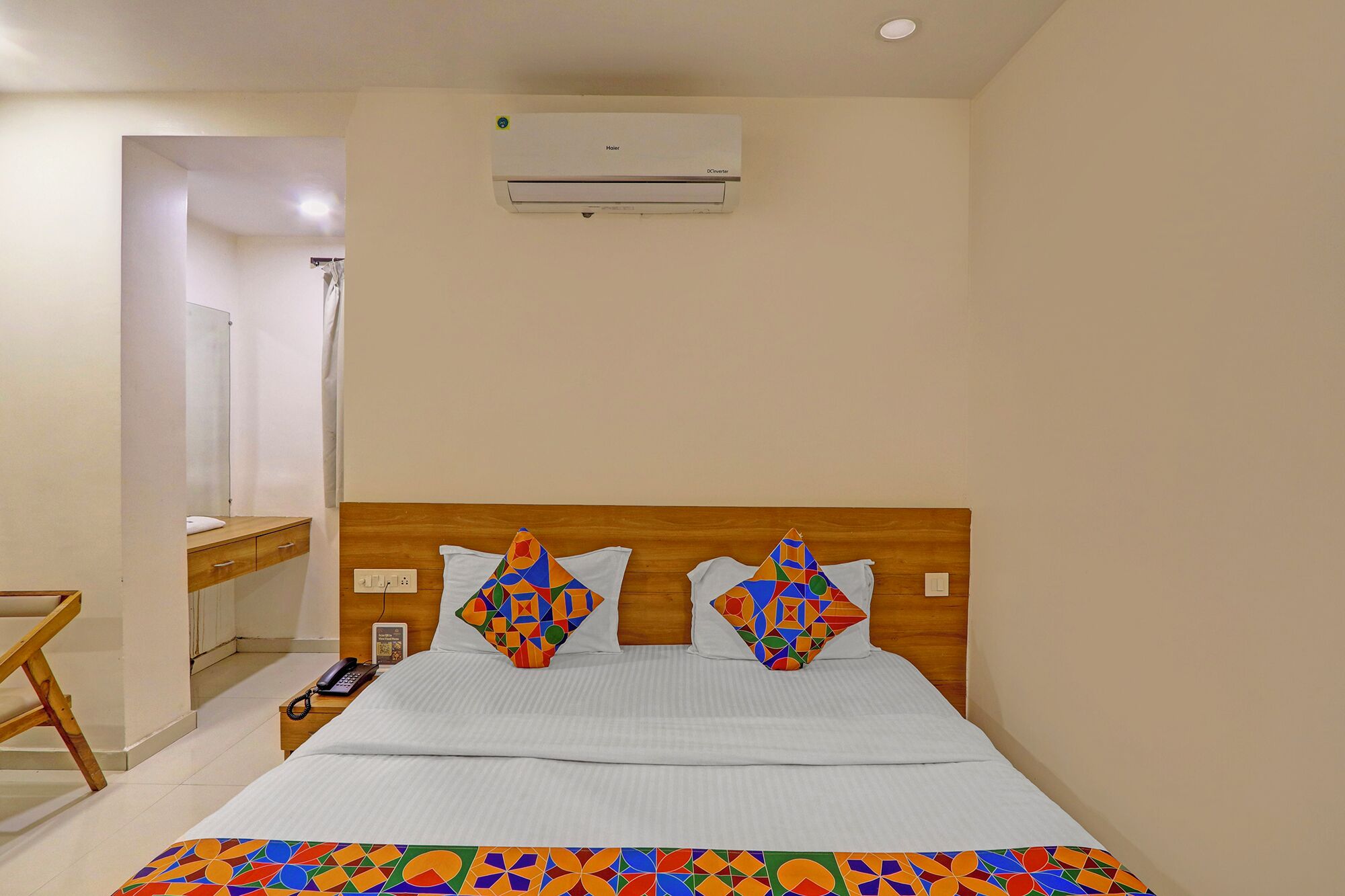 A hotel room with a wooden headboard, a bed with a colorful geometric-patterned bedspread, a bedside table with a phone and a book, and an air conditioning unit on the wall.
