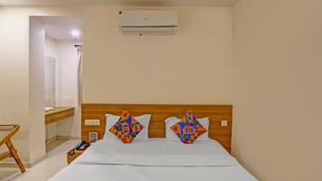 A hotel room with a wooden headboard, a bed with a colorful geometric-patterned bedspread, a bedside table with a phone and a book, and an air conditioning unit on the wall.