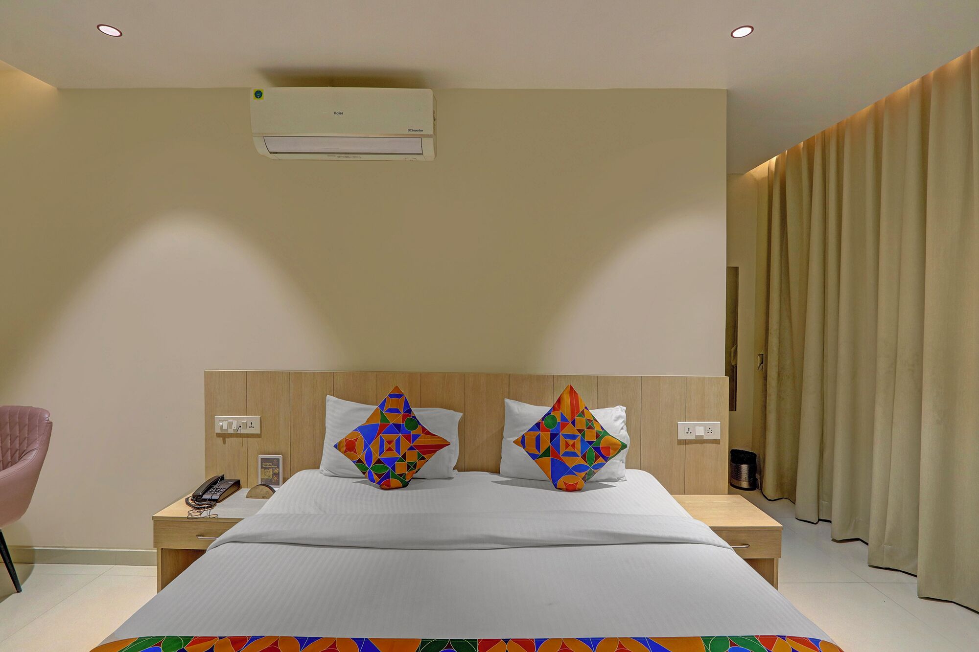 A hotel room with a bed, two pillows with geometric patterns, a bedside table with a phone, and an air conditioning unit on the wall.