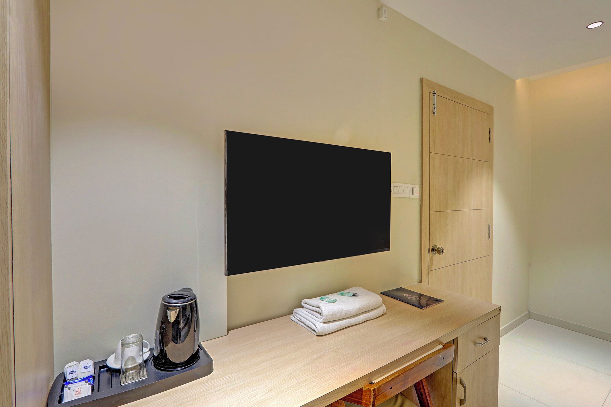 Ett hotellrum med en platt-TV som är monterad på väggen, ett skrivbord i trä med en vattenkokare, en hopvikt handduk och ett litet bricka.