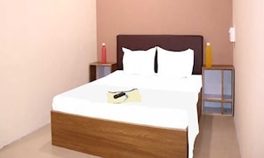A neatly made bed with a wooden headboard, a bedside table with a bottle, and a nightstand with a lamp. - Fabhotel Nova Rooms (Surat)
