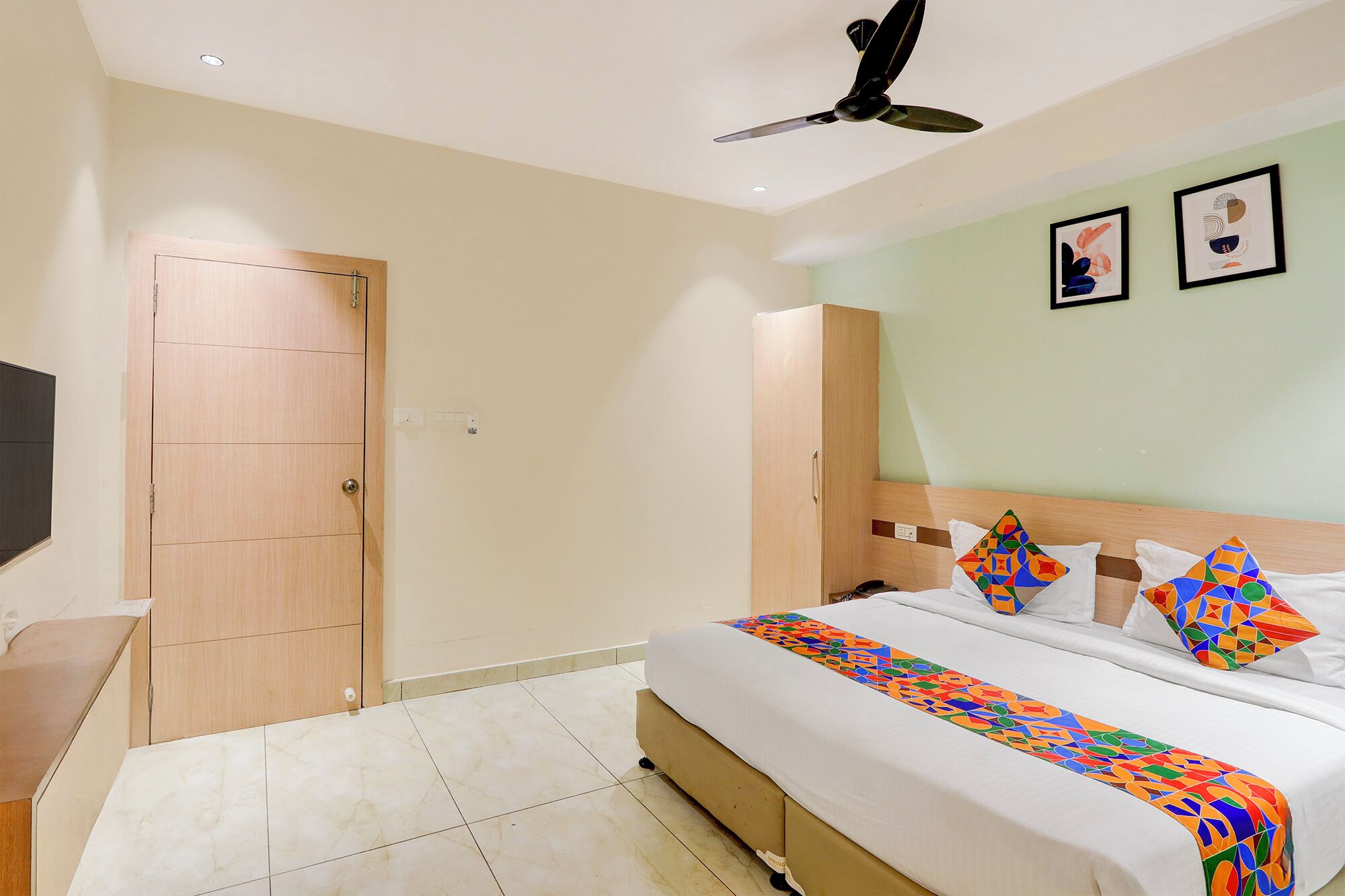Une chambre d’hôtel avec un lit, un ventilateur de plafond, une télévision et des œuvres d’art accrochées au mur.