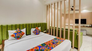 A modern hotel room with a green upholstered headboard, a bed with white bedding and colorful geometric pillows, a wooden partition, and a kitchenette area with a TV.
