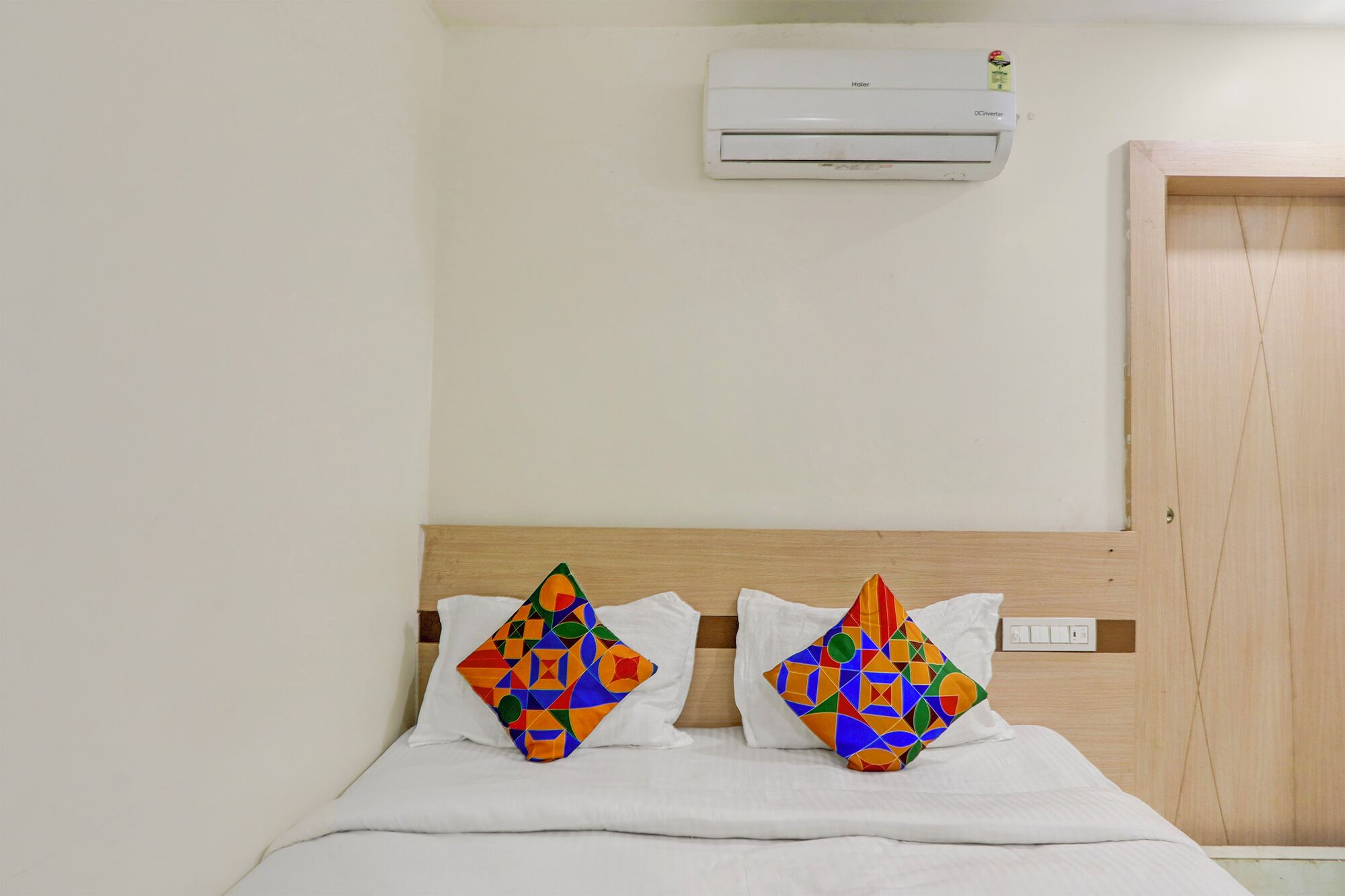 Habitación de hotel con cama, dos almohadas coloridas, cabecera de madera, pared blanca y aire acondicionado.
