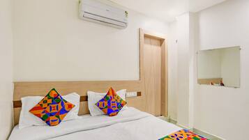 A hotel room with a wooden headboard, a bed with white bedding, colorful geometric pillows, an air conditioning unit, a wooden door, and a small mirror on the wall.