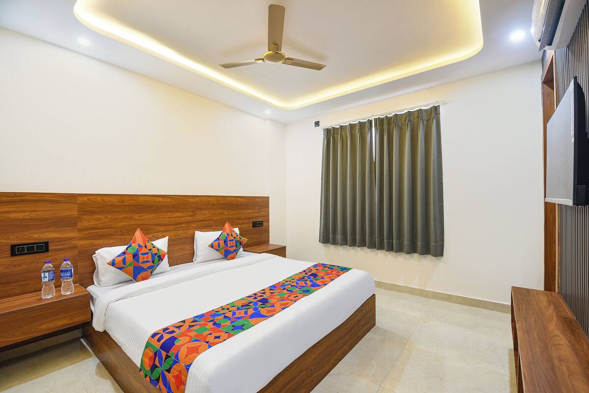 A hotel room with a wooden headboard, a bed with white bedding, colorful pillows, a ceiling fan, a window with curtains, and a flat-screen TV on the right side.