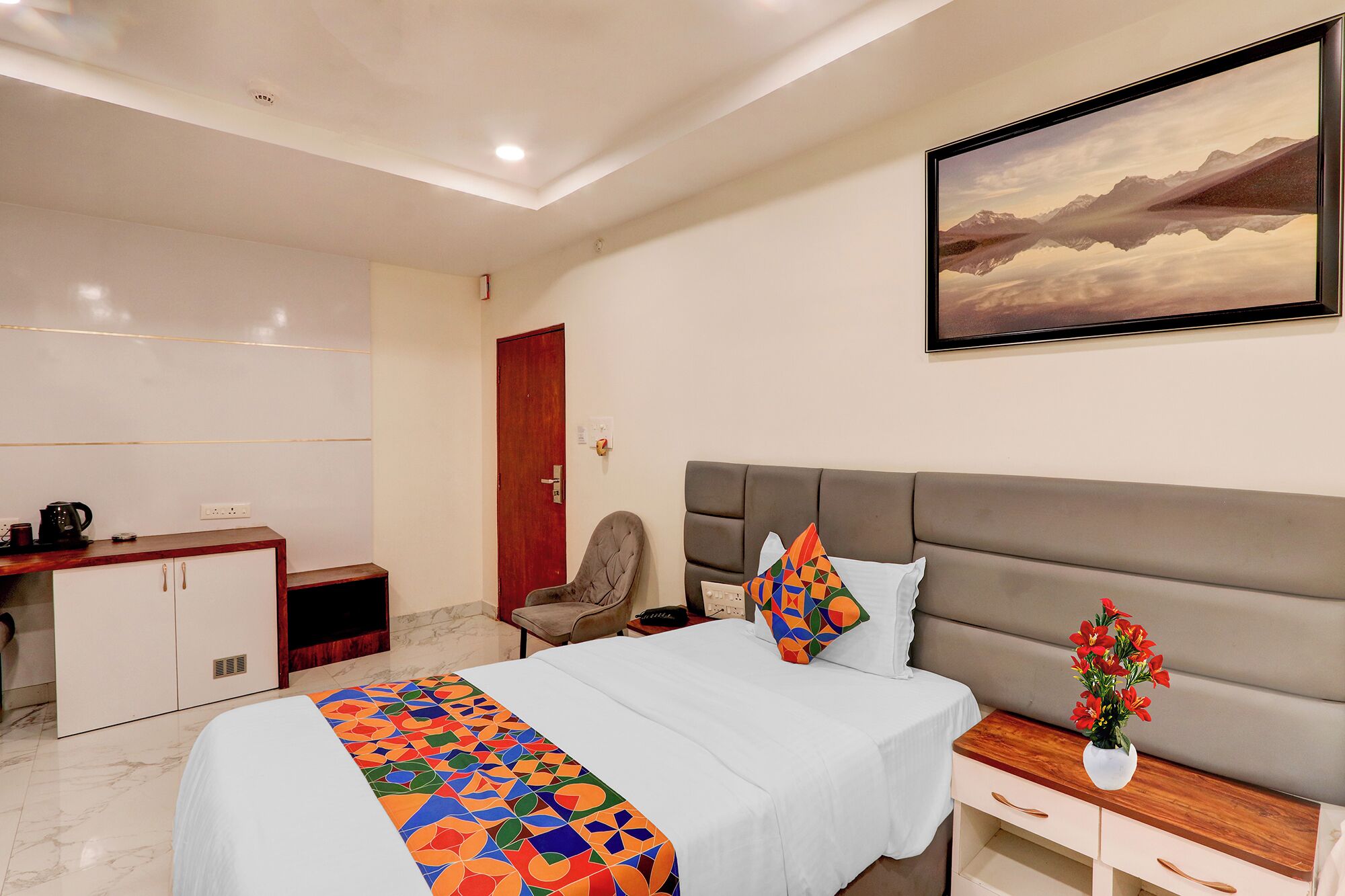 A hotel room with a bed, a desk, a chair, a nightstand, and a painting on the wall.