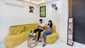 A yellow sofa with a small round table and a fan in a modern room with white walls and a ceiling light. - Fabhotel Radha Rani Residency (Khammam)