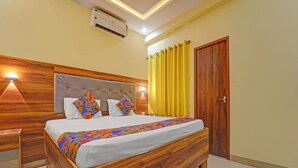 A bedroom with a wooden bed, a headboard, a nightstand, a wall-mounted air conditioner, and a yellow curtain. - Fabhotel Amutham Residency (Kanyakumari)