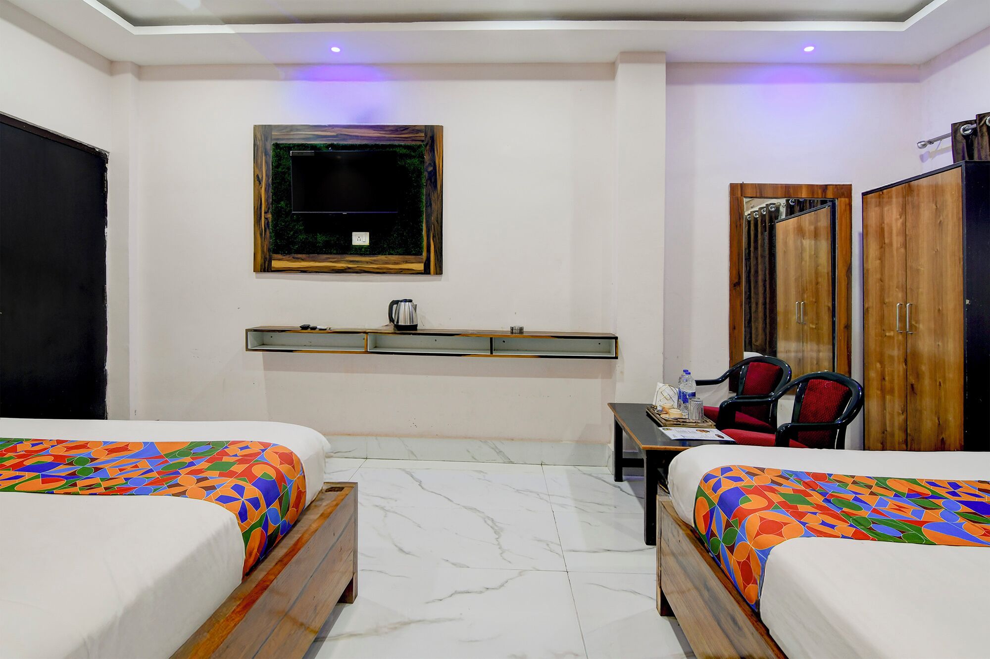 Habitación de hotel con dos camas, un escritorio con silla, un armario, un televisor y una estantería con hervidor.