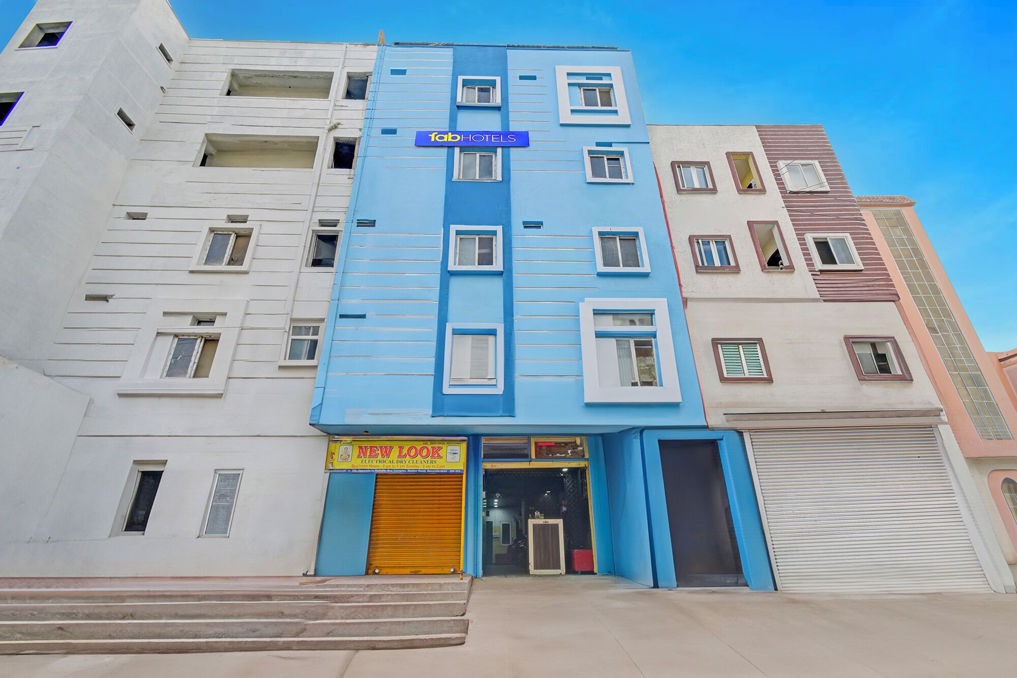 A multi-story hotel with a blue facade, a visible entrance, and a ground-level business with a yellow shutter.