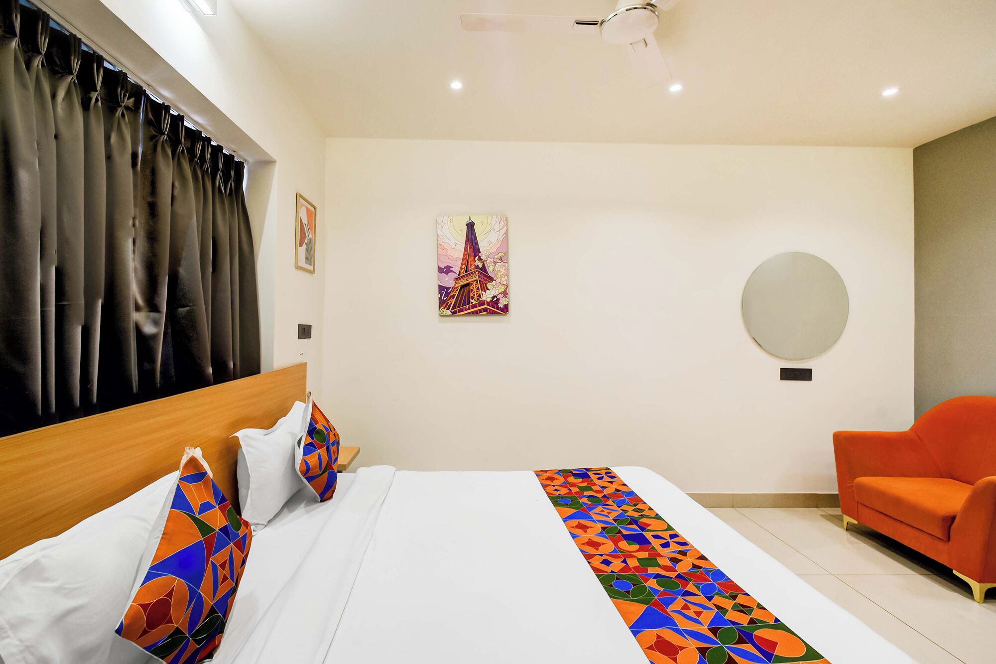 Una habitación de hotel con una cama, una silla, un cuadro y un espejo redondo en la pared.