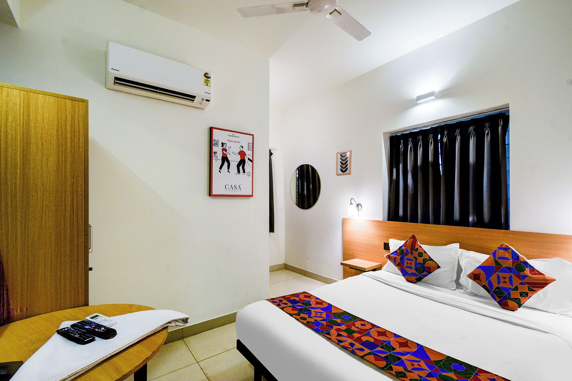 A hotel room with a bed, a nightstand, a desk, a chair, a ceiling fan, an air conditioner, a window with curtains, and a framed picture on the wall.