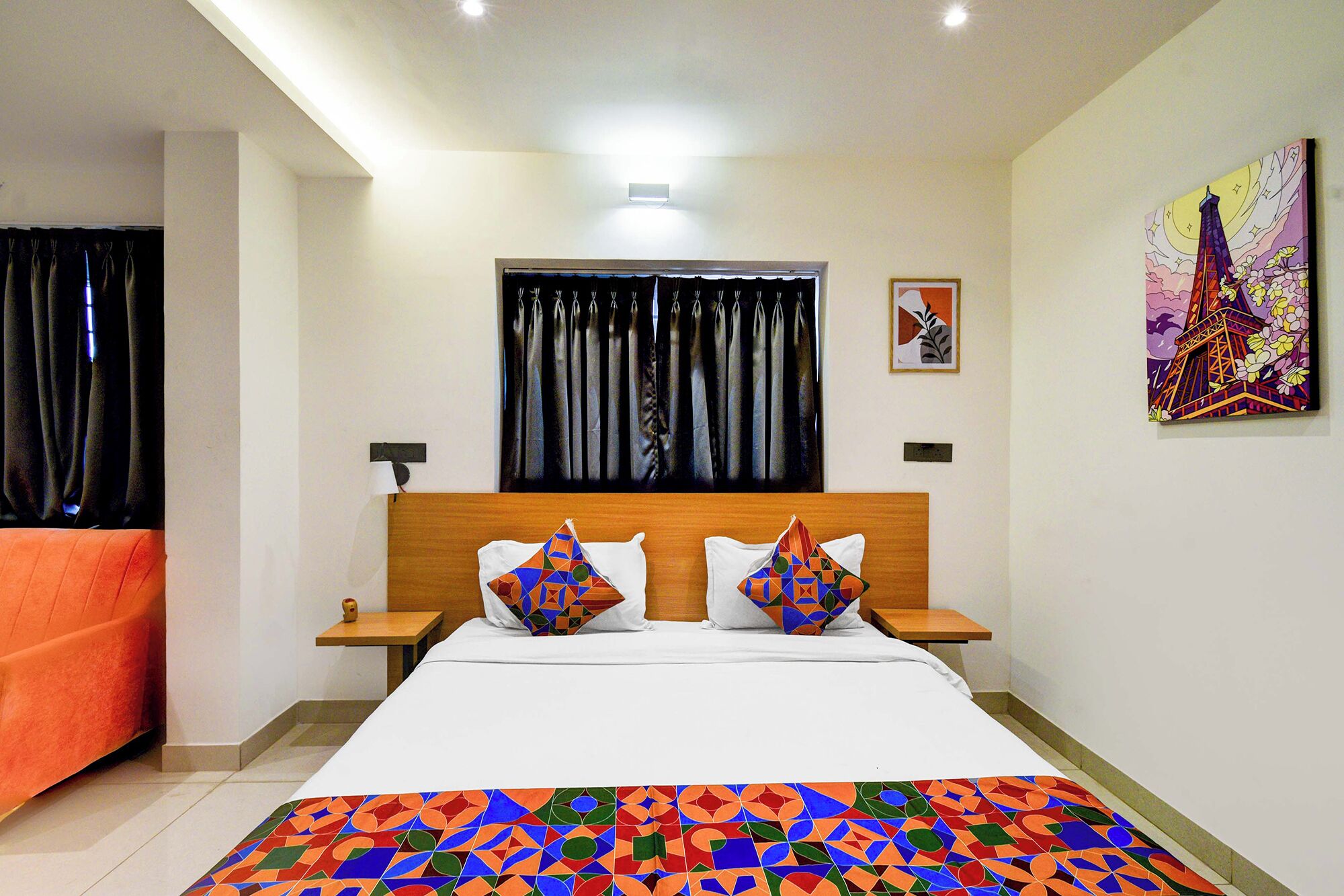 A hotel room with a bed, two nightstands, a wooden headboard, a window with black curtains, a colorful artwork on the wall, and an orange chair.