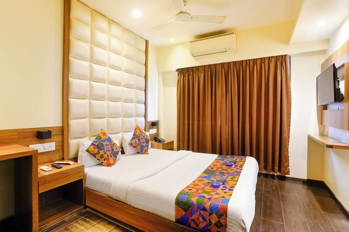 A hotel room with a large bed, a wooden nightstand, a desk, a ceiling fan, an air conditioning unit, and a TV on the wall.