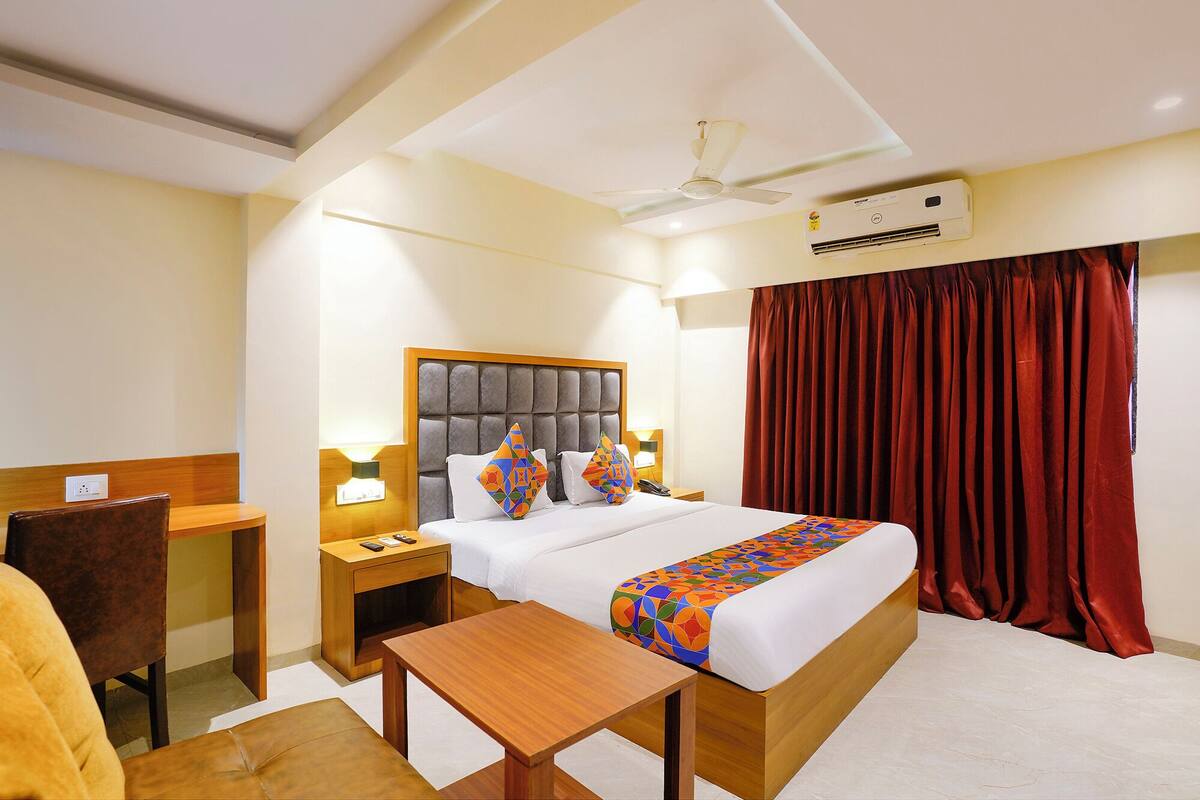 A hotel room with a bed, a desk, a chair, a nightstand, a table, a ceiling fan, and an air conditioning unit.