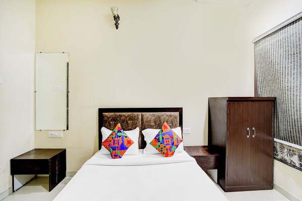 Un dormitorio con una cama, dos almohadones geométricos y coloridos, un armario de madera, una mesita de noche y una lámpara fijada a la pared.