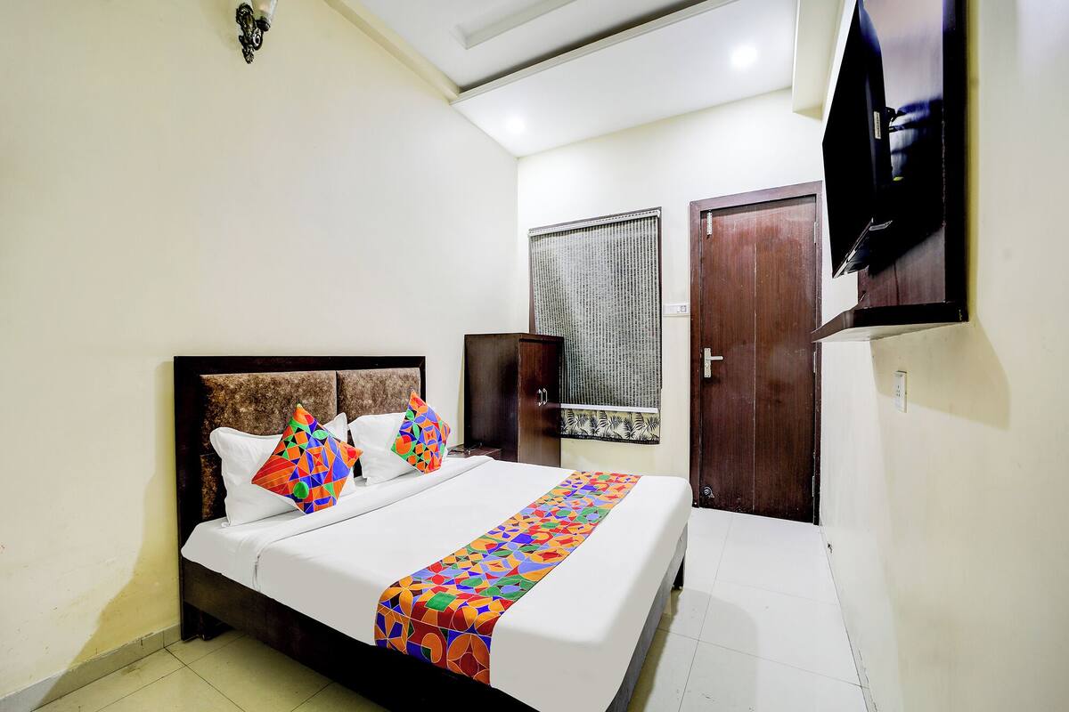A neatly made bed with colorful pillows and a patterned bedspread, a wooden wardrobe, a television mounted on the wall, and a window with curtains.