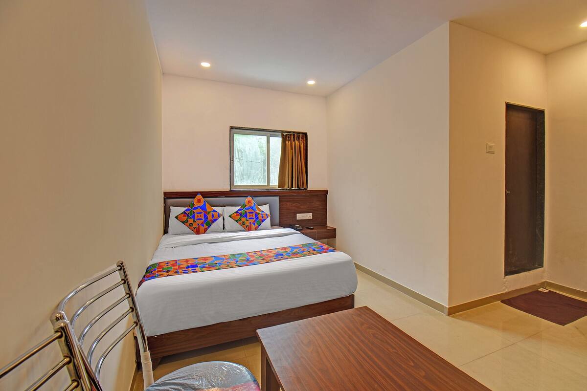 A hotel room with a bed, a wooden nightstand, a window with curtains, a door, and a small wooden table.