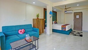 A hotel room with a blue sofa, a bed with a colorful blanket, a wooden wardrobe, a small table with water bottles, and a ceiling fan. - Fabhotel Small Daddy Plus (Calangute)