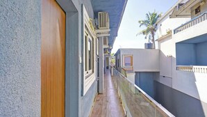 A narrow corridor with a wooden door on the left, a balcony with a glass railing on the right, and a blue building with air conditioning units. - Fabhotel Small Daddy Plus (Calangute)