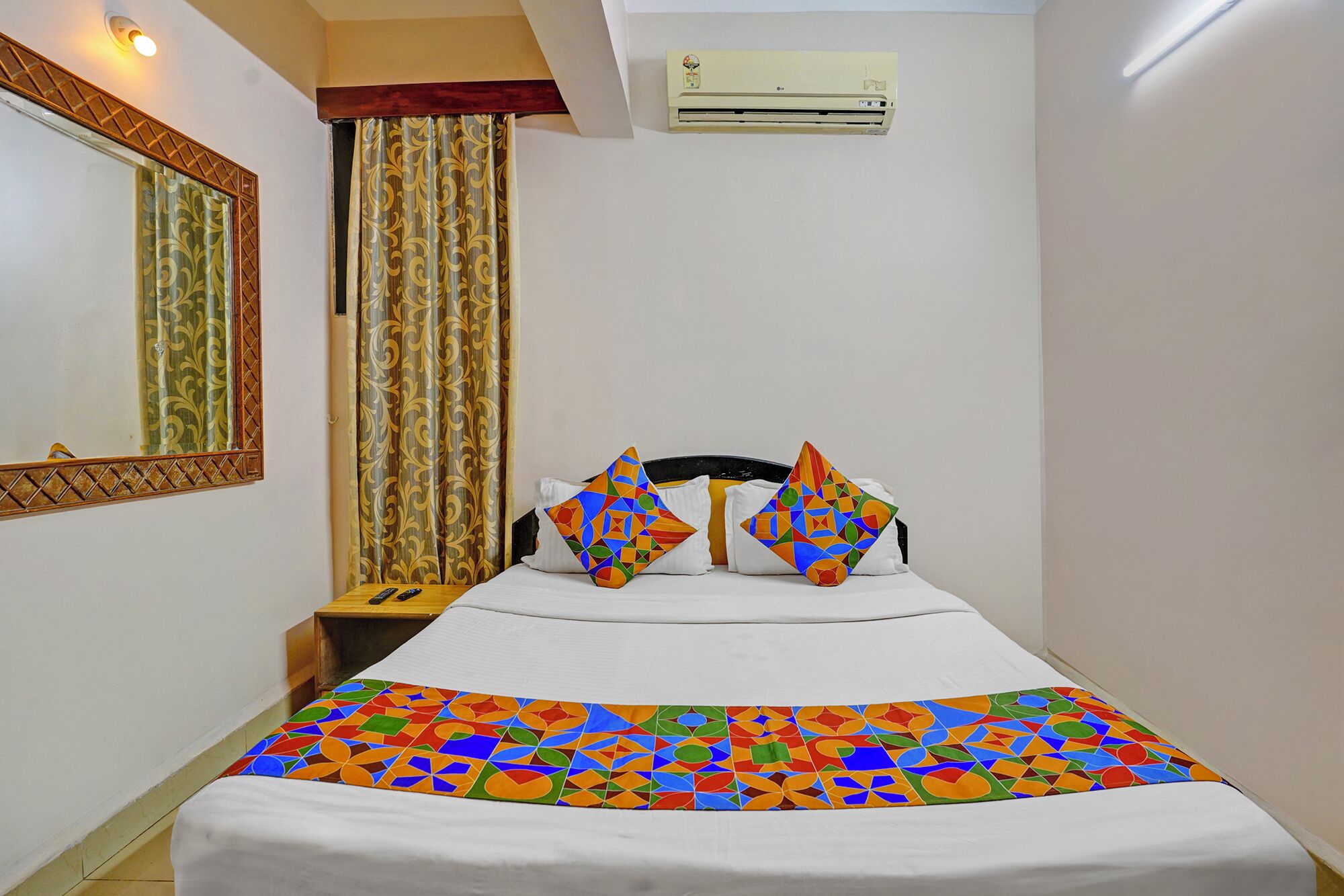 Habitación de hotel con cama, dos almohadas, mesita de noche, espejo, cortinas y aire acondicionado.