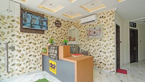 A hotel reception area with a wooden reception desk, a clock, a TV mounted on the wall, and a patterned wallpaper. - FabHotel Aurika Retreat (Indore)
