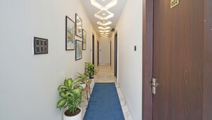 A hallway with a blue carpet, potted plants, framed artwork, and a wooden door with a number plate. - FabHotel Aurika Retreat (Indore)