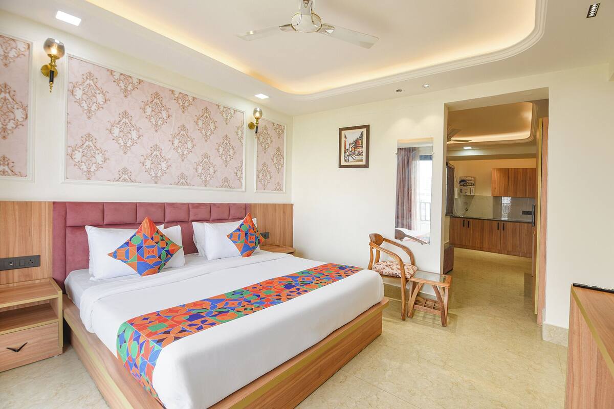 A hotel room with a bed, bedside table, and a small desk with a chair. There is a kitchenette in the adjacent room.