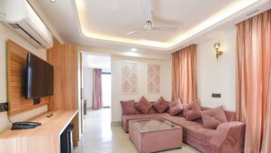 A modern living room with a sectional sofa, a glass coffee table, a flat-screen TV mounted on a wooden cabinet, and a ceiling fan. - Fabhotel Prime Atithyam (Gurugram)