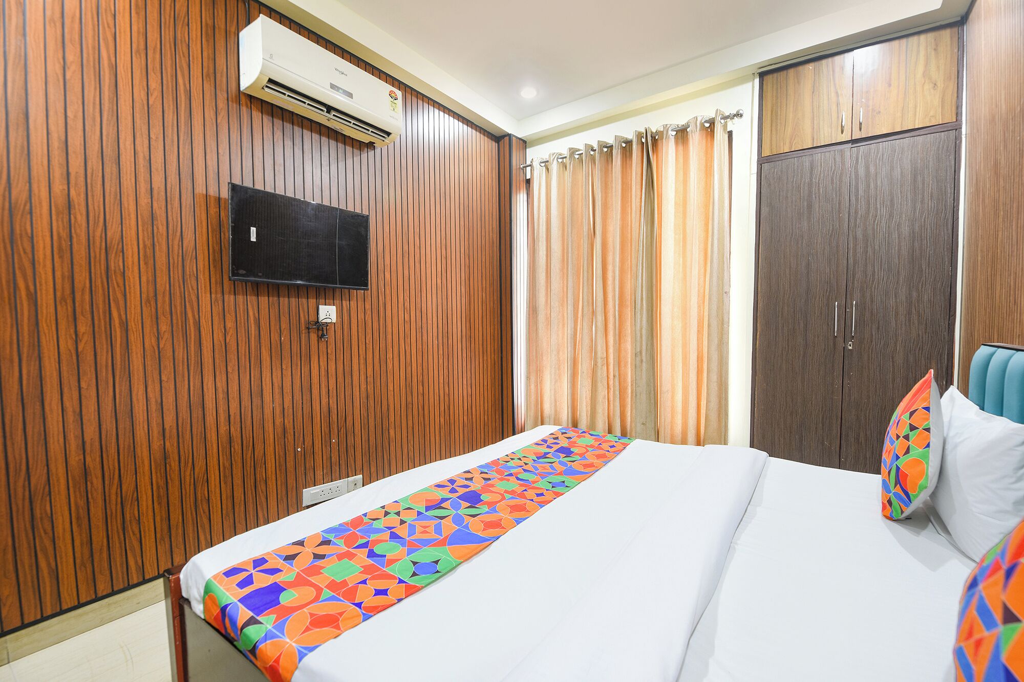 A hotel room with a bed, a TV mounted on the wall, wooden paneling, a wardrobe, and a curtain.