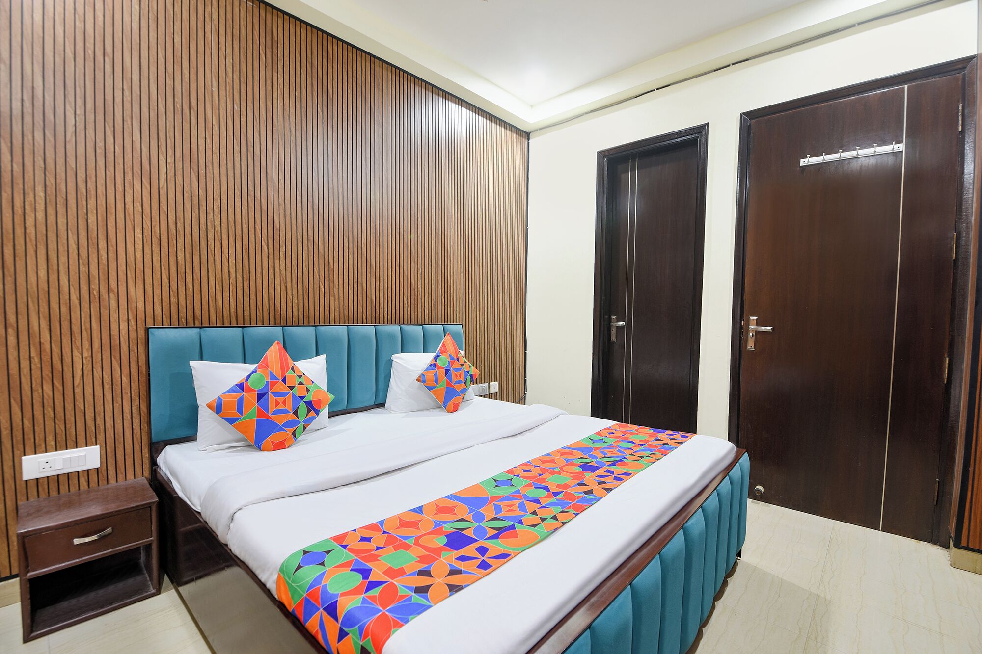 A hotel room with a wooden panel wall, a blue upholstered headboard, a bed with white bedding and colorful geometric pillows, a nightstand, and a wardrobe.
