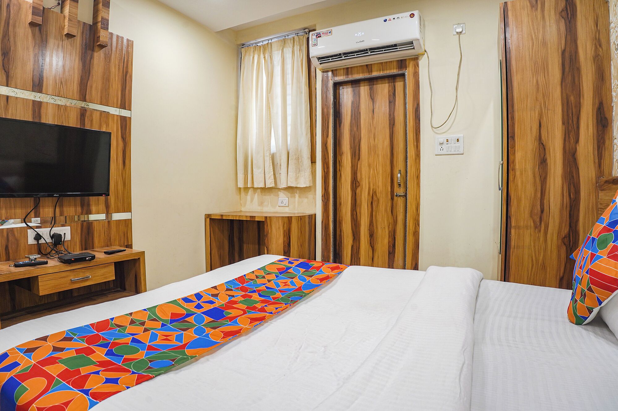 A hotel room with a bed, a TV, a wooden wardrobe, and an air conditioning unit.