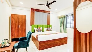 A bedroom with a wooden wardrobe, a bed with colorful pillows, a blue chair, a desk with a kettle, and a ceiling fan. - Fabhotel Leon Square (Bengaluru)