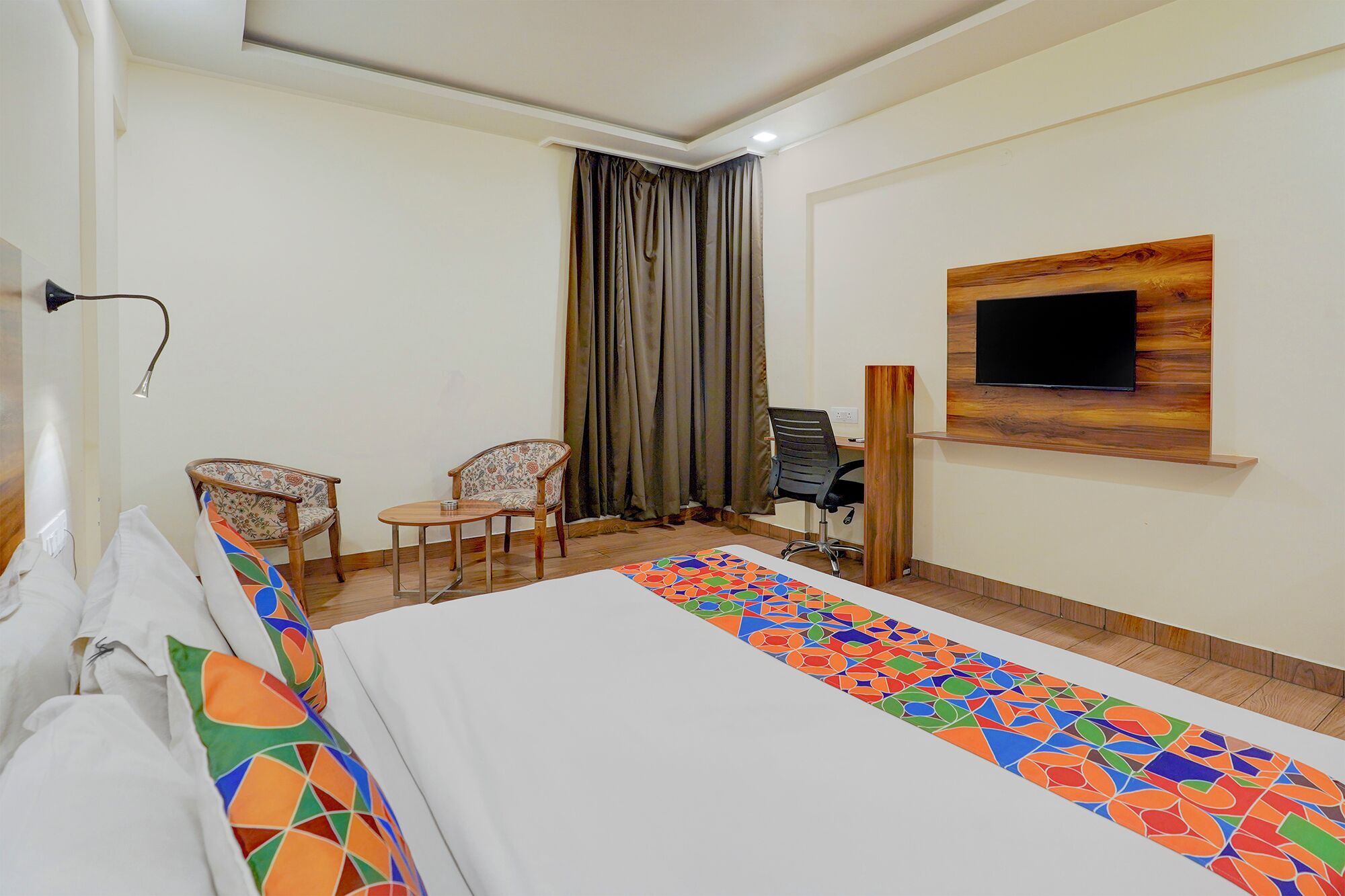 A hotel room with a bed, two chairs, a desk, a TV mounted on a wooden panel, and a window with curtains.