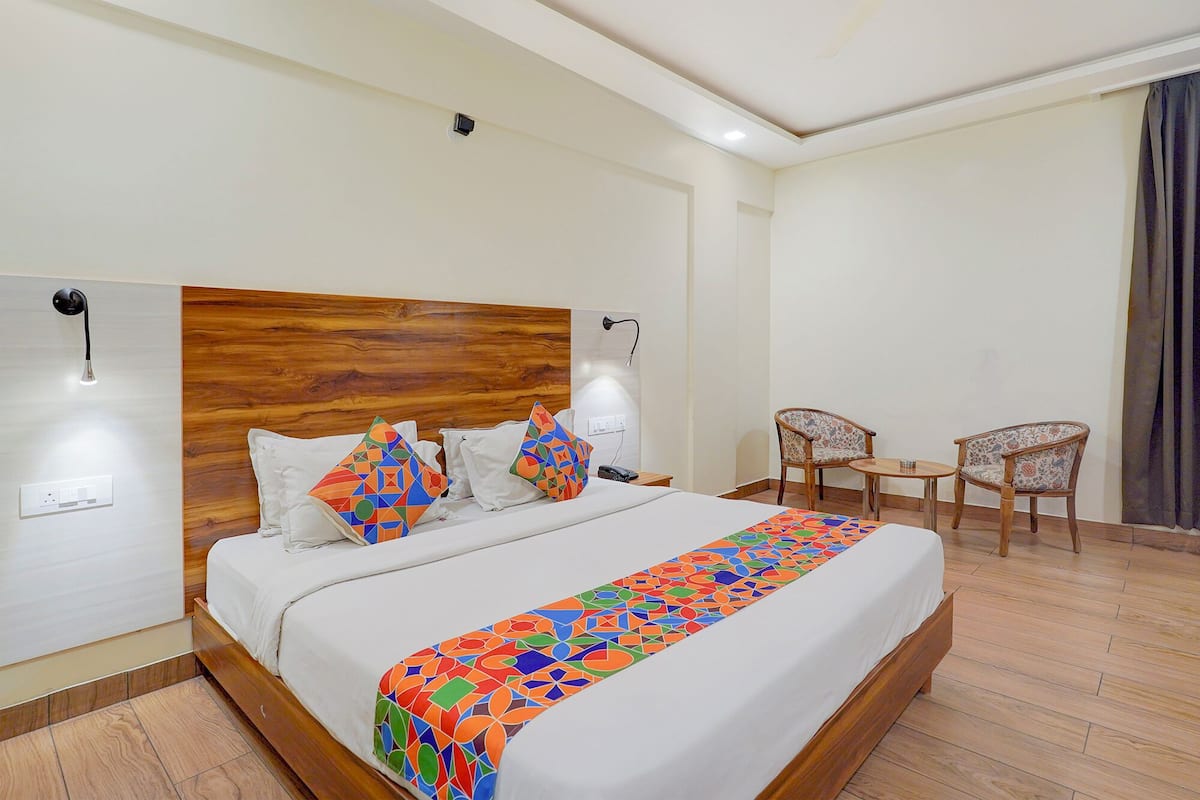 A hotel room with a wooden headboard, a bed with white linens, colorful geometric pillows, a bedside table with a lamp, two chairs, and a small table.