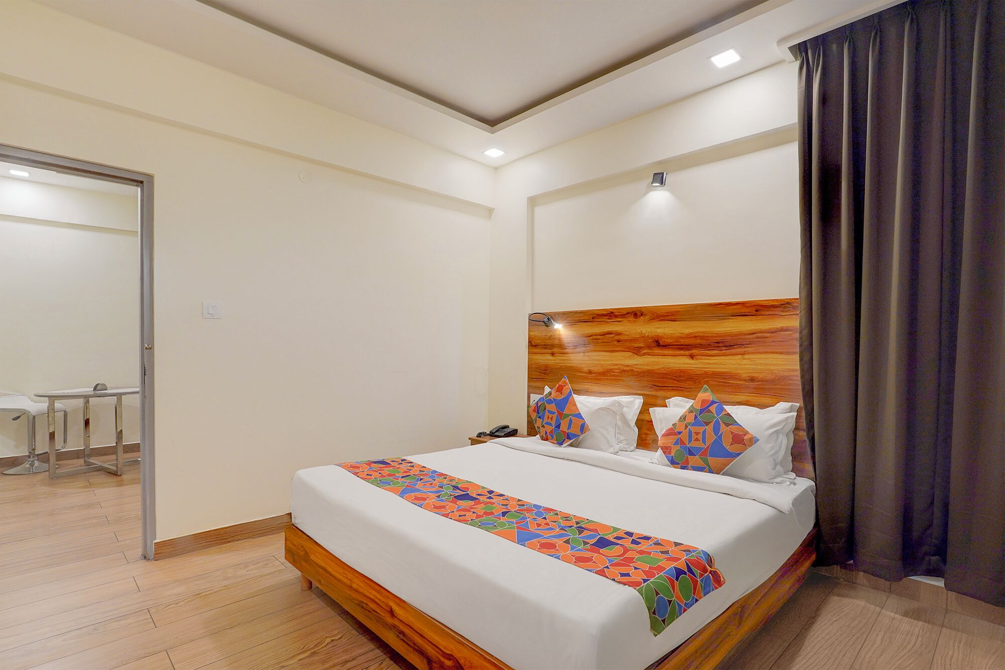 A hotel room with a wooden headboard, a bed with white bedding, colorful pillows, and a small bedside table.
