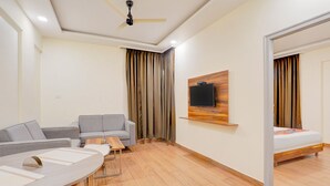 Habitación de hotel moderna con sofá, mesa de comedor, televisor montado en un panel de madera y cama con estructura de madera.