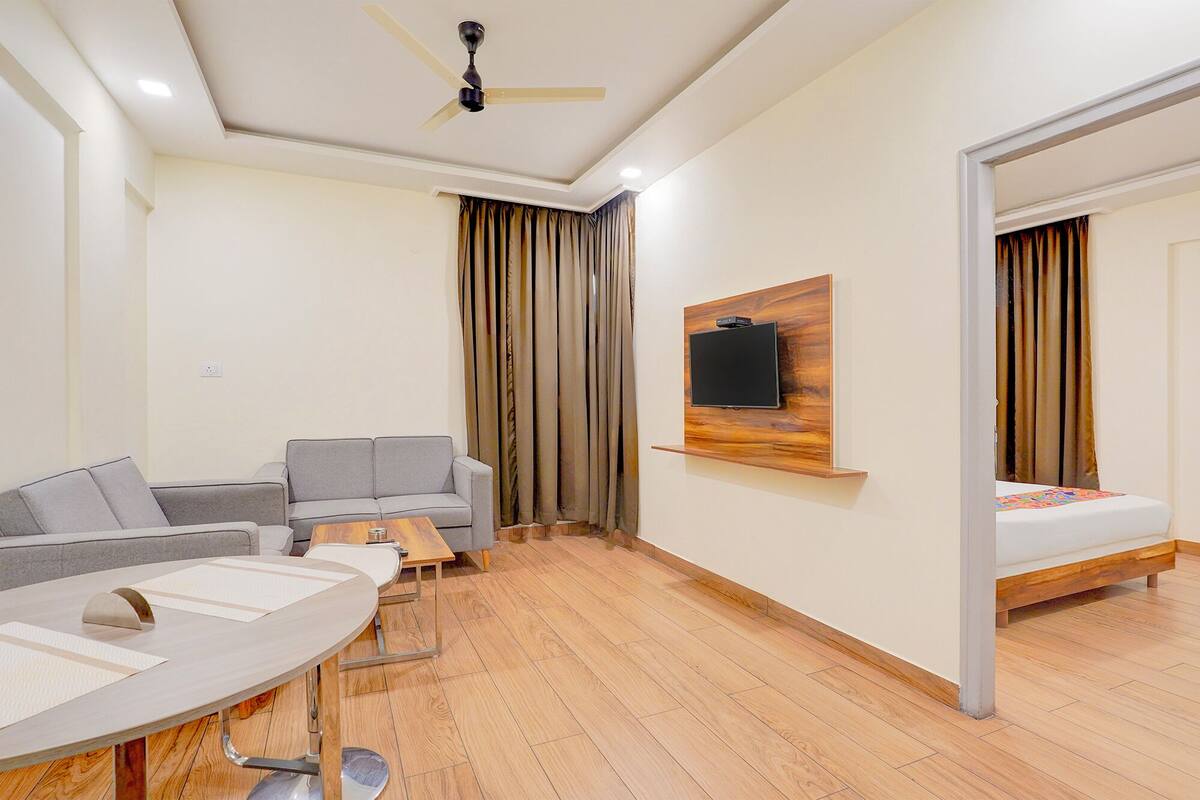 A modern hotel room with a sofa, a dining table, a TV mounted on a wooden panel, and a bed with a wooden frame.